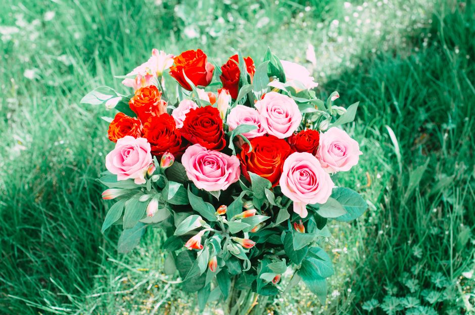 A vibrant bouquet of fresh flowers featuring pink roses, bright red roses, and orange-tipped blossoms arranged together and wrapped in light pink paper with a ribbon. The bouquet is held upright again