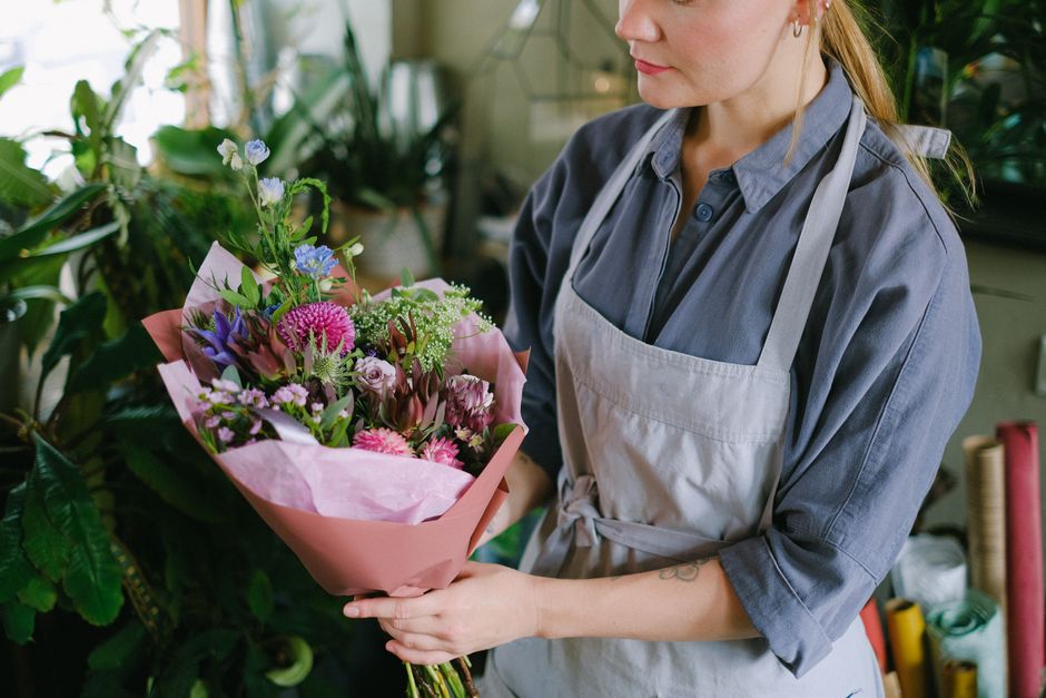 A person wearing a blue button-up shirt and a light gray apron holds a vibrant floral bouquet in both hands, positioned near the chest. The bouquet features a mix of fresh flowers with various texture