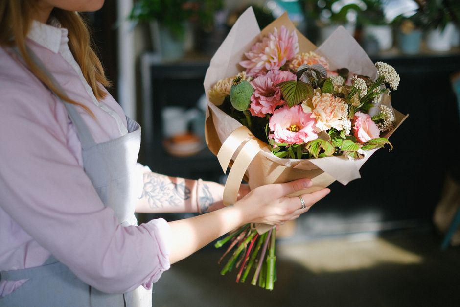 A person with long, light brown hair wearing a light pink shirt and a gray apron is holding a fresh floral bouquet wrapped in light brown paper with a cream-colored ribbon. The bouquet features pink a