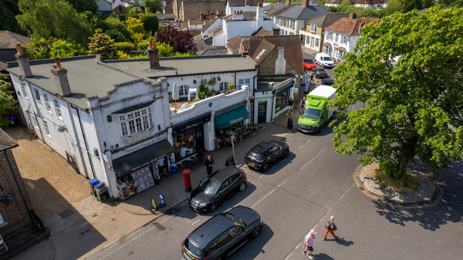 An aerial view of a small, white, two-story building on Kenton High Street, hosting a flower shop with a blue awning. The shop displays floral arrangements near the entrance. Parked in front are sever