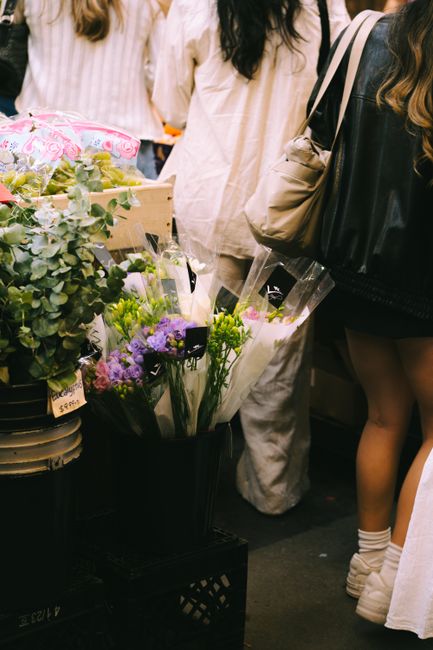 A bouquet arrangement of fresh, vibrant purple and green flowers, wrapped in clear cellophane with black tags, is being held by a person at a flower market. Surrounding the bouquet are other floral di