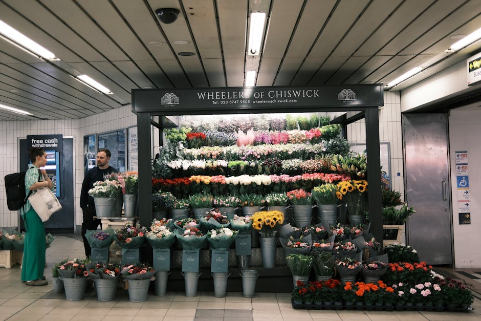 A vibrant floral display at Wheeler of Chiswick in a busy underground station. The arrangement features a variety of fresh flowers including white, pink, red, yellow, and purple blooms, carefully arra