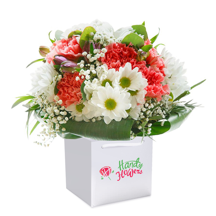 Bursting with freshness and joyous colour, this professionally arranged bouquet is displayed upright in a sharply-designed white square gift bag, creating a striking focal point against a seamless white background. The artistry of the bouquet shines through coral and peach carnations, whose ruffled, voluminous petals deliver a visual feast, setting a warm and welcoming tone. Clusters of crisp white daisies with striking lime-green centres punctuate the display, their pure brightness reminiscent of the lively gardens found in the Kenton Road area. Wispy white baby's breath interlaces between the larger blooms, bringing softness and movement, while glimpses of closed purple-tipped buds introduce intrigue and dimension. Encircling the flowers, glossy, deep green leaves form an elegant, natural collar-impeccably arranged to hold the bouquet's shape and exude sophistication. The white gift bag, decked with sturdy ribbon handles, is not only stylish but also practical for both shopside pick-up and doorstep delivery, capturing the spirit of convenience that Kenton locals value. The light is soft and diffused, lifting every shade and detail for ultimate clarity. This image exemplifies the expert care and floral creativity of Kenton's florists and is the perfect visual for any article celebrating the ease and comfort of sending thoughtfully curated flowers-be it for birthdays, noteworthy occasions, or just because.