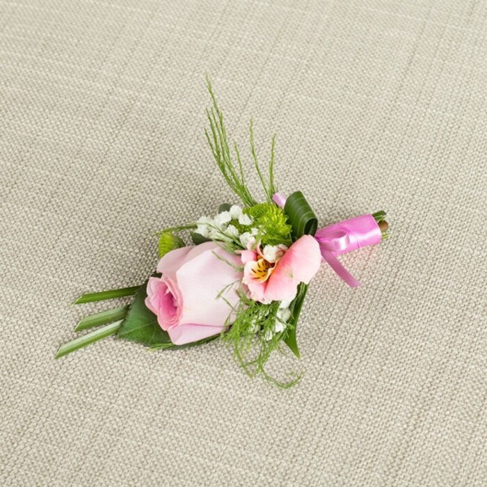 Lying diagonally atop a neutral, delicately textured linen fabric reminiscent of Kenton's refined homes, this handsomely arranged boutonniere-or wrist corsage-embodies care and contemporary artistry. The key highlight is a pastel pink rosebud, tightly ruffled yet just beginning to unfurl, symbolising tenderness and new beginnings. Surrounding the rose, fine sprays of pure white baby's breath bring airiness and old-world charm, interspersed with miniature green cushion mums that contribute a refreshing vibrancy. A soft, peachy-pink bloom-perhaps an Alstroemeria-opens nearby, its centre kissed by yellow and fine markings for unique visual depth. The foliage structure is artfully composed: fern-like sprigs and spiky, slender greens fan ever so slightly upward, evoking a wild freshness found along the edges of Kenton Recreation Ground, while broader, deep green leaves-some folded-offer contrast and support at the base. A wide dark green leaf spirals neatly to bind the stems, finished with a striking fuchsia pink satin ribbon knotted in a meticulous bow. Bathed in gentle light that accentuates the contrasting textures and subtle blush hues, this accessory is both festive and refined-a fitting choice for weddings, formal events, or significant life milestones celebrated in and around Kenton.